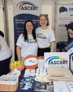 Two women who are ASCERT staff members smile for a photo. They are wearing white t-shits with a rainbow coloured ASCERT logo. They stand in front of an ASCERT roller banner and an ASCERT merchandise table.