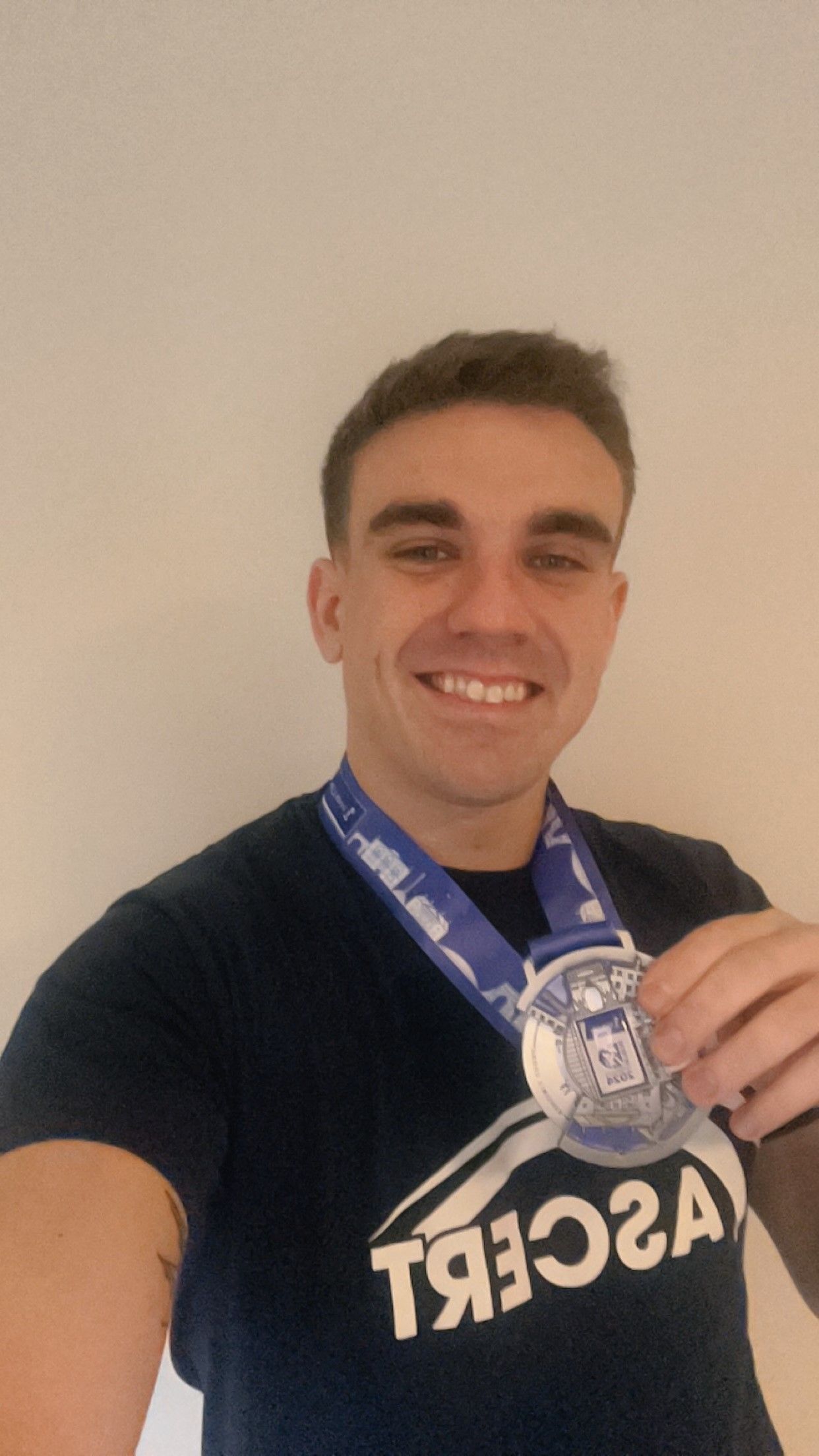 Martin is a young man in his 20s with brown hair. He wears a navy ASCERT t-shirt and smiles at the camera while holding up his Dublin Marathon Medal.