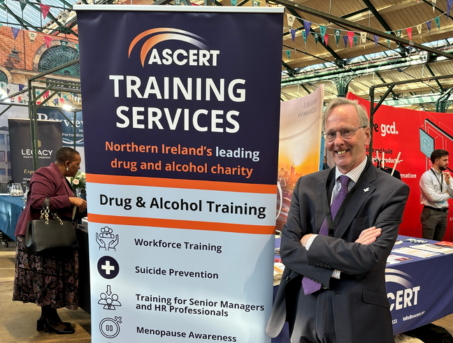 Colin, ASCERT's Training Services Manager, is wearing a suit. He has his arms folded and is smiling next to a roll up banner outlining ASCERT's Training Services.