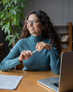 A woman with long, brown, curly hair, wearing glasses holds a pen in both hands and looks smiling over her shoulder. She is sitting at a desk with her notebook and laptop in front of her.